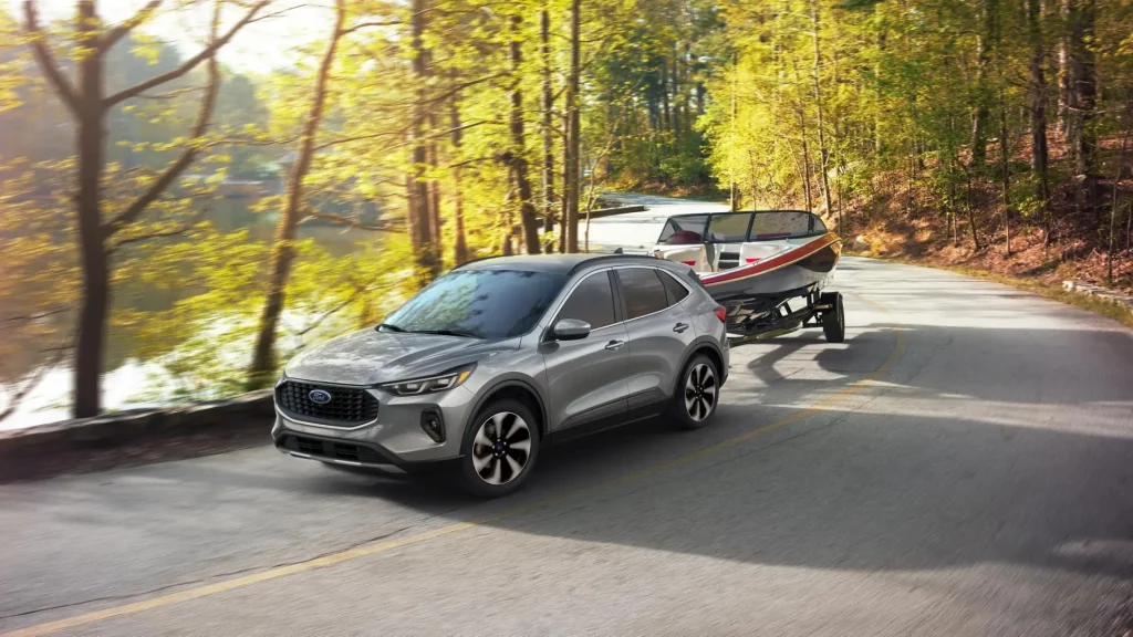 A silver 2024 Ford Escape SUV towing a boat on a scenic road surrounded by trees.