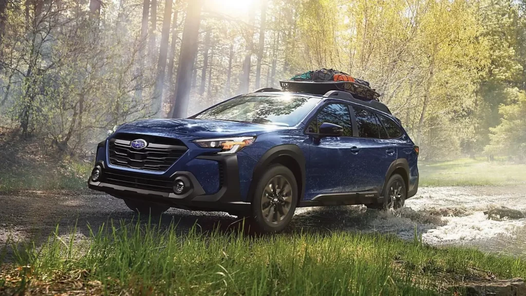 A blue 2024 Subaru Outback driving through a forested area with a roof rack loaded with camping gear.